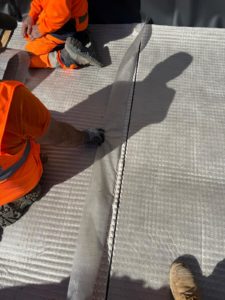 BATI-IDF deux compagnons en gilets orange en train de rabouter deux lés de nappe drainante en pied de façade, savoir-faire BATI-IDF — Boissy-sans-Avoir (78).