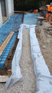 BATI-IDF vue longue du drain bordé de cailloux drainants enveloppés dans le géotextile, le long du pavillon, équipe BATI-IDF en arrière-plan — Ouges (21).